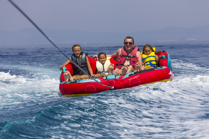 A family enjoying a tubing ride on the sea, holding tightly onto an inflatable tube as a speedboat pulls them across the water, with waves splashing around them.