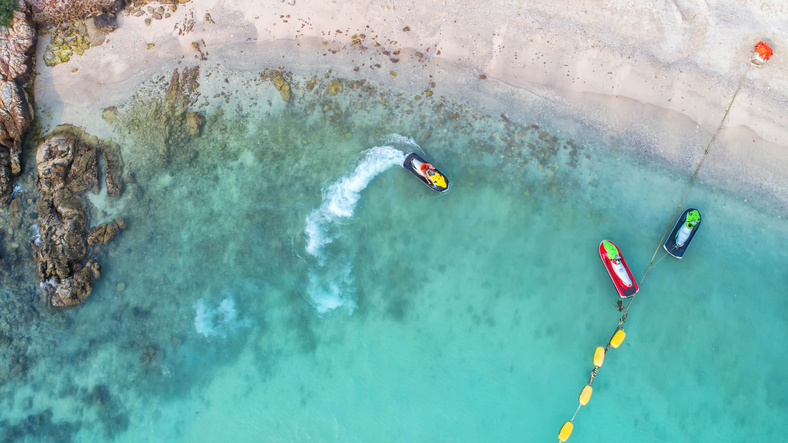 Aerial view of several people riding jet skis across the open sea, leaving white foamy trails behind them on the deep blue water.
