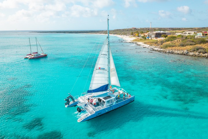 Catamaran sailing on clear turquoise water near a sandy shore under a partly cloudy sky.