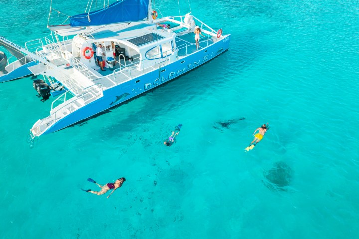 Catamaran on clear water with three snorkelers swimming nearby.